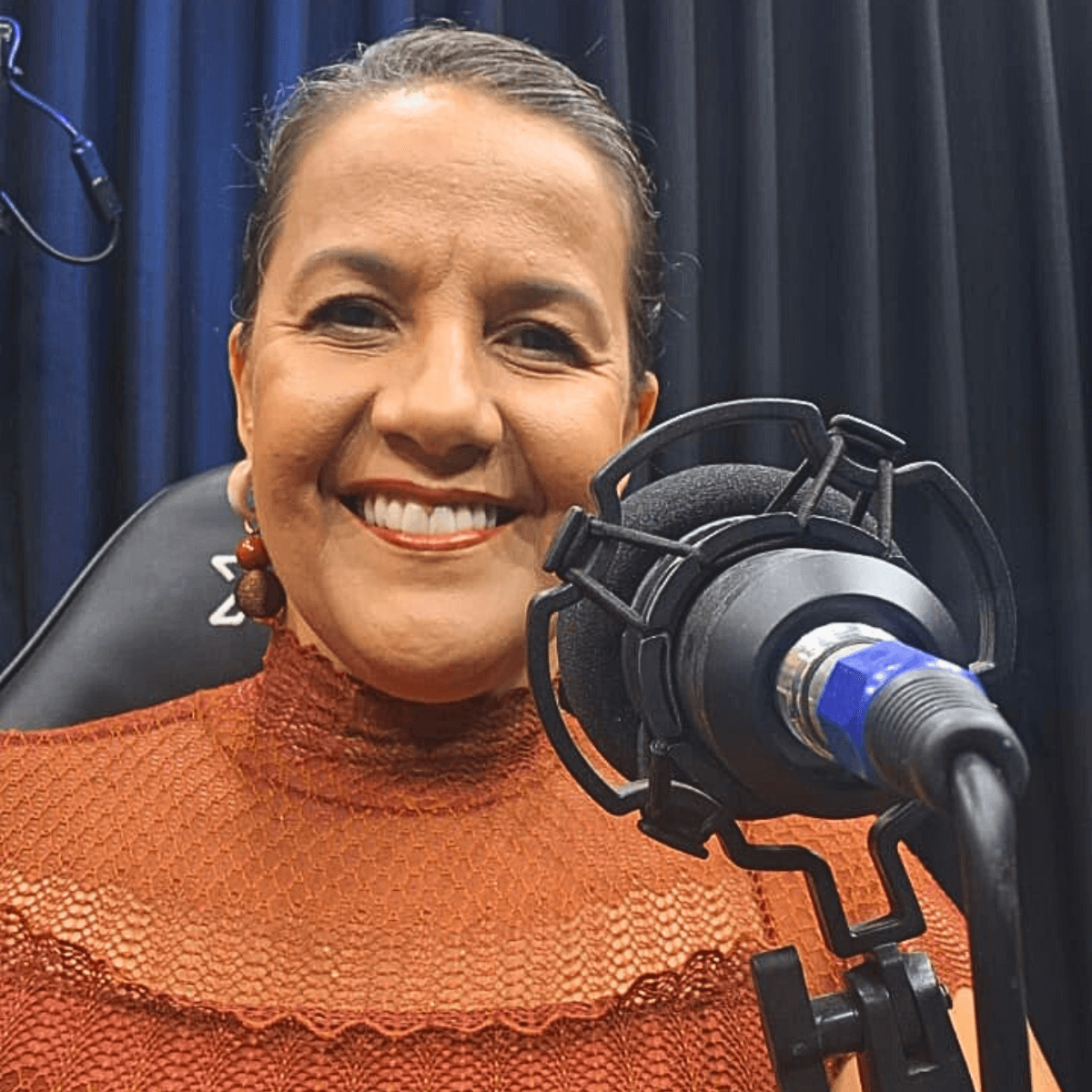 Close de Lili Machado sorrindo enquanto fala em um microfone de podcast.