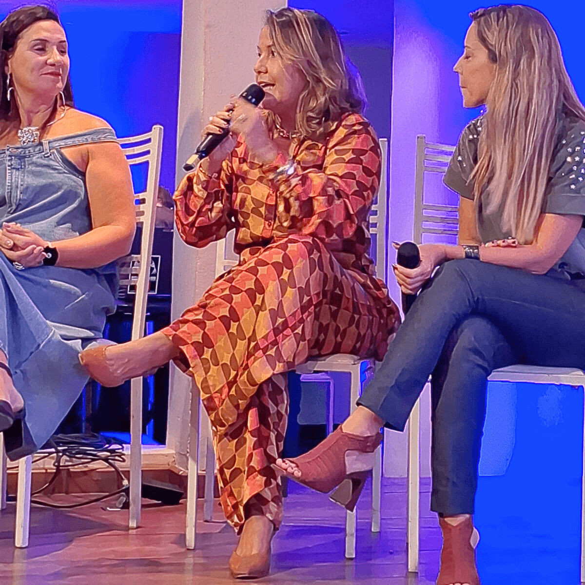 Lili Machado sentada em um banco alto no palco com outras duas mulheres, segurando um microfone.