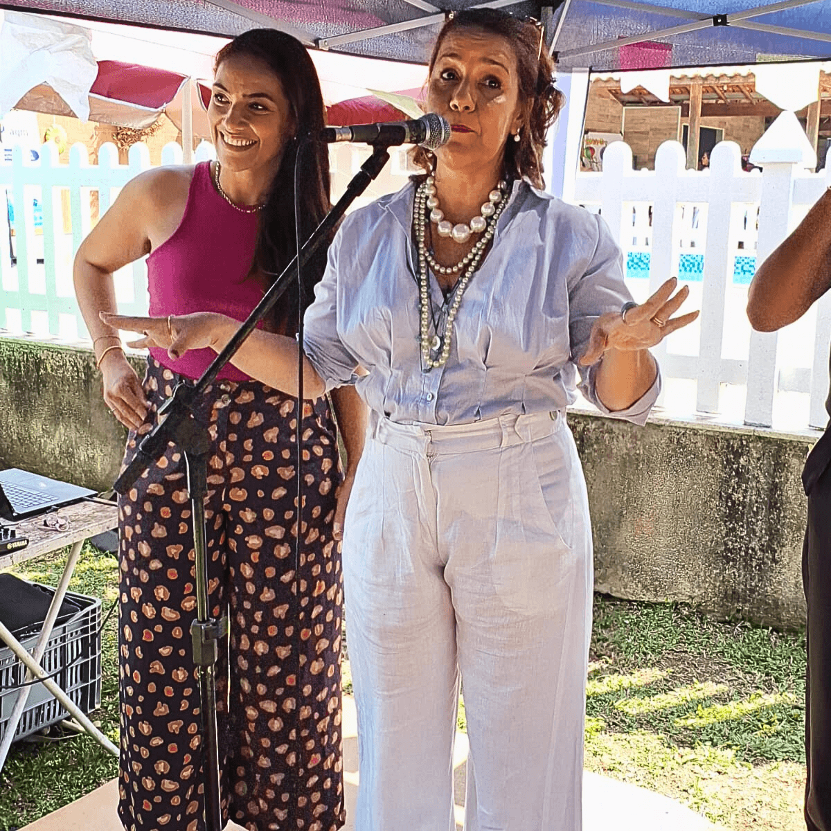 Duas mulheres em pé, lado a lado, uma segurando um microfone em um evento.
