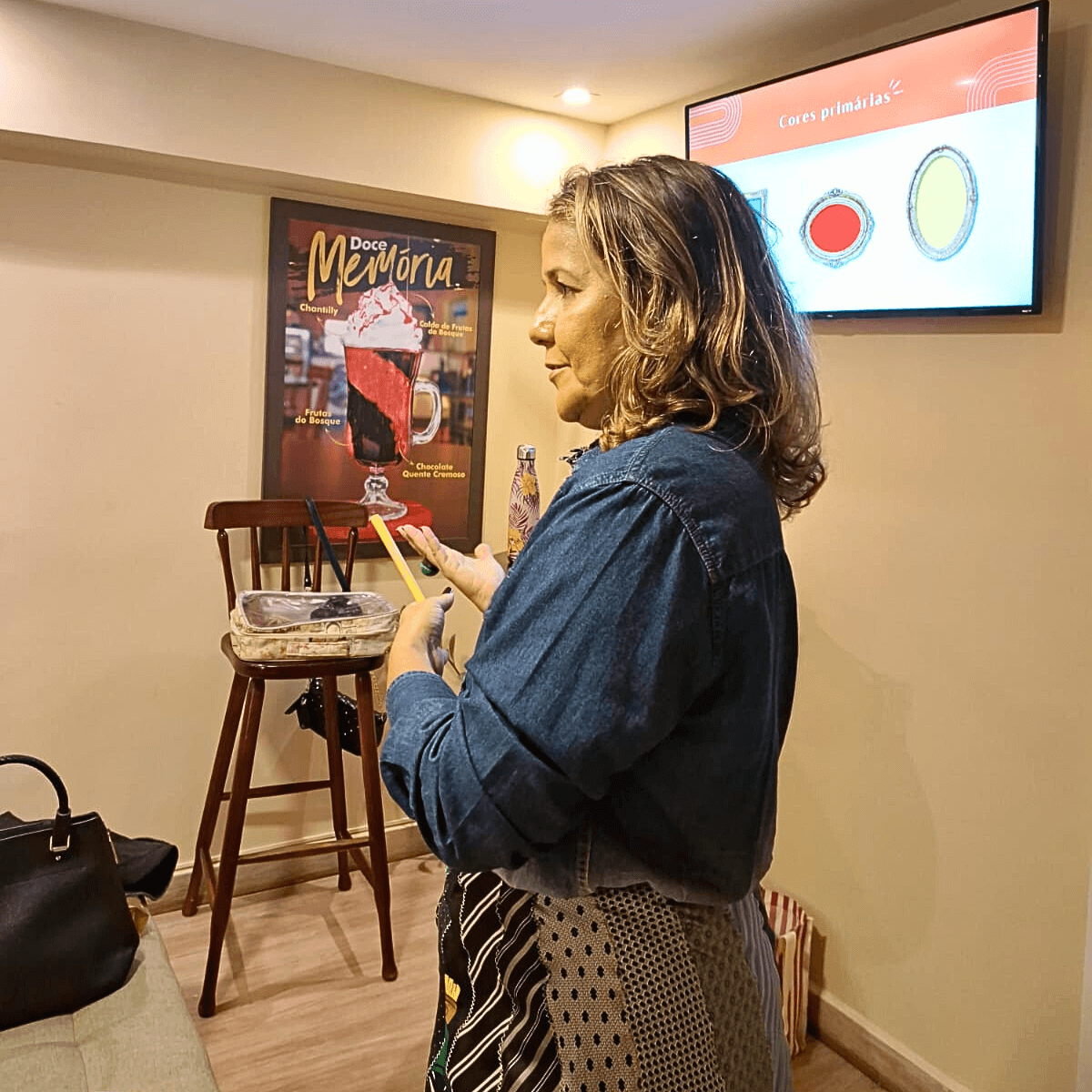 Lili Machado em pé em um ambiente interno, olhando para o celular ou um objeto pequeno.