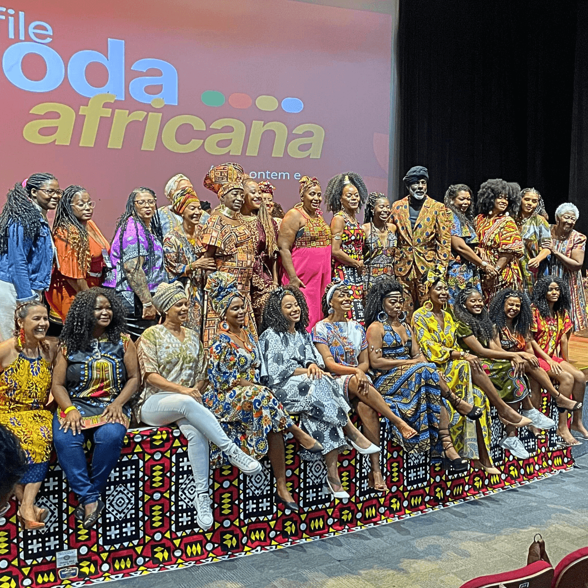 Grande grupo de mulheres sentadas e em pé em uma arquibancada, com uma apresentação na tela ao fundo.
